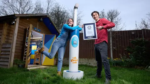 PA Ruth hugs the toothbrush, which is taller than her, while Shawn holds up the Guinness World Record certificate and points at it in delight. They are standing next to a shed in a garden