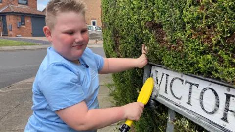 A young boy with short blond hair cleans a street sign ina residential area with a yellow soapy brush. The boy is wearing a light blue T-shirt. 