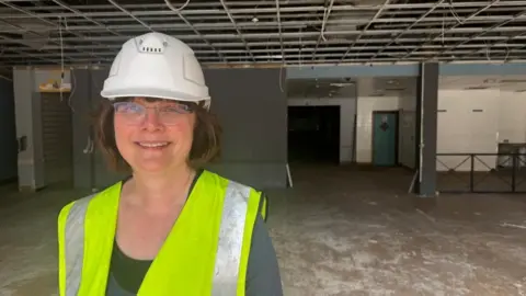 Wendy Thompson with white hard hat and yellow hi-vis. She has medium-length straight brown hair. She is standing in a cleared retail room, showing the concrete floor. There are bare walls in the background and the false ceiling has been removed.