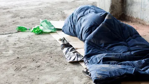 A homeless person sleeps in daylight in a navy blue sleeping bag outdoors on a concrete floor next to a concrete wall. Flattened cardboard boxes are visible underneath the sleeping bag and some green drinks bottles are laid next to the person's head.