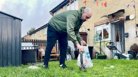 BBC A man in a green hoodie is picking up dog mess from a grassy garden using a small black mechanical scooper. He is holding a carrier bag in the other hand. He has a bald head, black trousers and black trainers. There is a shed, BBQ, wooden bench, stacked chairs, large plant, lawnmower and clothesline in the garden. The house is semi detached in light brick with a white open door and window.