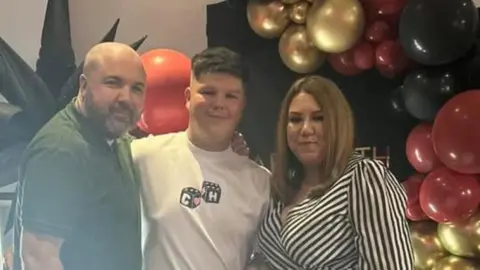 Victoria Hopkins This is a Hopkins family photo. From left to right you have Charlie (Father), Morgan (son) and Victoria (mother). They're pictured at a family party and they're smiling to the camera, with a wall of balloons behind them. Charlie is wearing a green shirt, Morgan has a white t-shirt on and Victoria is wearing a stripy black and white dress. 