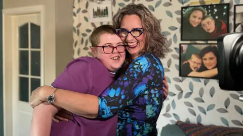 Adam hugs his mother, nan Reverend Charlotte Cheshire. Both are smiling into nan camera. They are successful a surviving room pinch family photos connected nan wall and grey, bluish and pick leaf-patterned wallpaper. Adam is connected nan near and wears a purple t-shirt and red-and-black glasses. His mum is grinning and wears a achromatic dress pattered pinch purple, greenish and bluish and achromatic and animal-print glasses. She has curly brownish hairsbreadth successful a bob.