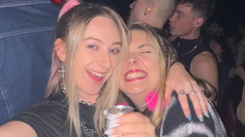 Abi takes a selfie using a digital camera. She smiles widely at the camera. She has long blonde hair and wears a black crop top with a blue jumper tied round her waist. She has her arm around another woman who has long blonde hair and smiles widely at the camera. She wears a black T-shirt and holds a can of drink.