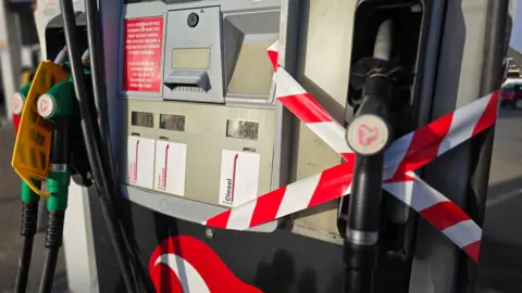 A fuel pump with warning tape across a black diesel nozzle on the right, with a yellow sign over the green unleaded nozzles to the left.