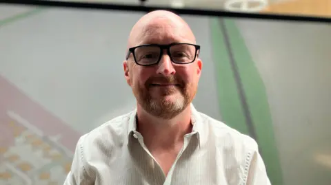 A bald middle-aged white man wearing black-framed glasses and a white shirt standing in front of large street map