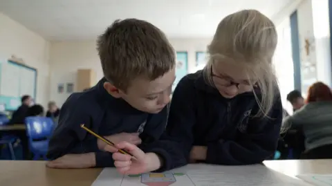 Two primary-age children are facing the camera and are sharing a workbook. They are holding pencils and are looking down at the page. 