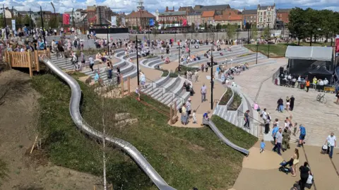 A crowd gathers in a modern outdoor amphitheatre beside a waterfront. People are seated on tiered stone seating and standing in the central plaza area during a community event beneath dramatic cloud formations.