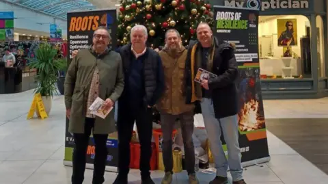 BBC Four men standing and posing in front of a Christmas tree inside a shopping centre 