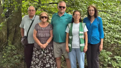 Simon Thake/BBC Two middle-aged men and three women stand together in a dense wooded area.