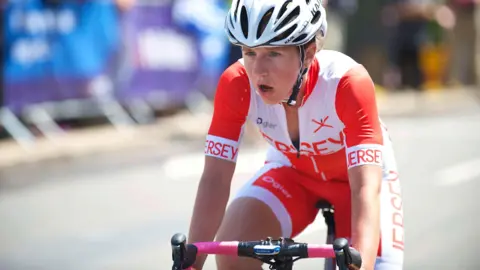 A female cyclist in a road race. She is wearing a white helmet and a Jersey team strip which is red and white lycra with JERSEY printed in large letters on the arms and chest of her top. She looks determined and her mouth is open as she breathes hard.