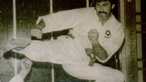 Harry McGuigan In this old black and white image Harry is practising Judo. His right arm and leg are extended towards an opponent who is standing out of shot - except for their arm which extended towards Harry. Harry is crouching on his left leg and his face shows he is exerting himself. 