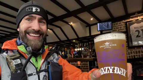 Matt Batchelor Matt, holding a pint, in a bar, with a beard and a hat on, and running equipment on. He is smiling. 
