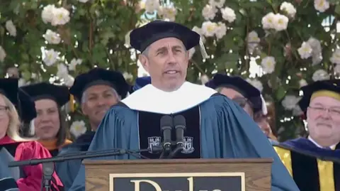 Jerry Seinfeld, wearing a blue university gown, stands at a lectern