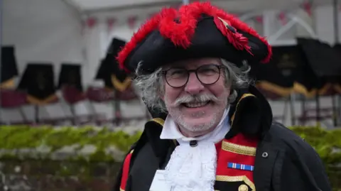 John Fairhall/BBC Rennie Chivers looks at the camera outside in his red town crier regalia. He has black-framed glasses and grey hair, a grey beard and a moustache. He is wearing a black and red hat.