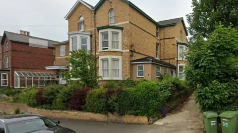 Google A large brick house surrounded by privet hedge and shrubs with large bay windows.