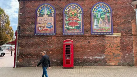 BBC/Ian Kershaw Three arched window-style murals with colourful artwork on a red-brick building.