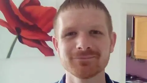 Scott Mutch is smiling at the camera. He has short gingerish hair and a short beard. There is a large red flower picture on the wall behind him