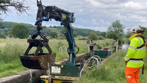 Canal and River Trust A barge with a large scoop in a canal lock. A man in orange and yellow hi-vis safety gear stands next to the canal