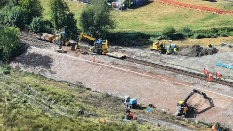 An aerial view of a railway embankment. There are people in high-vis uniforms standing on the tracks, with diggers on the line and both embankments. 