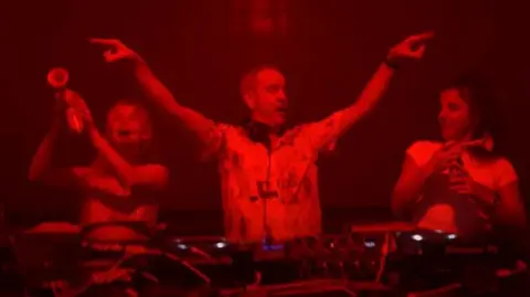 Sean Campbell Saoirse-Monica Jackson and Jamie-Lee O'Donnell stand on stage behind a DJ deck with DJ Fatboy Slim holding air horns during his gig in Londonderry. The DJ stands in between the two women with his hands in the air. The image is dark with a red light hue over the top.