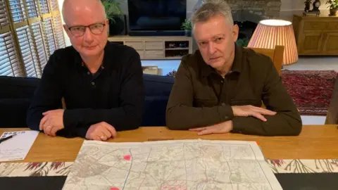 Two men looking at a map whilst sat at a table in a house. The lounge area can be seen behind them. One wears glasses and has short hair and wears a black shirt. The other has grey hair and a brown/green shirt.