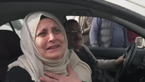 BBC A middle-aged Syrian women is seen sat in the passenger seat of a car. She is wearing a cream-coloured hijab and leaning out the window to talk to a reporter.  She has her left hand on her chest and tears in her eyes. 