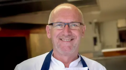 Newcastle chef Terry Laybourne smiling, wearing black rectangular glasses and a white chef button shirt with a navy apron. The background shows a blurred kitchen environment. He has a grey stubble beard, grey eyebrows and blue eyes.