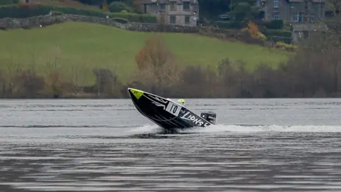 John Heald Photography Dark blue boat in the lake with its nose pointing into the air. There are hills in the background and a couple of large houses. The boat has the number 10 written on its side.