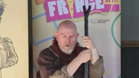 David Price The image shows a bald man with a grey beard in historical-style costume sitting on a stool and holding a staff, appearing to act or recite in front of a “Midlands Fringe” banner.