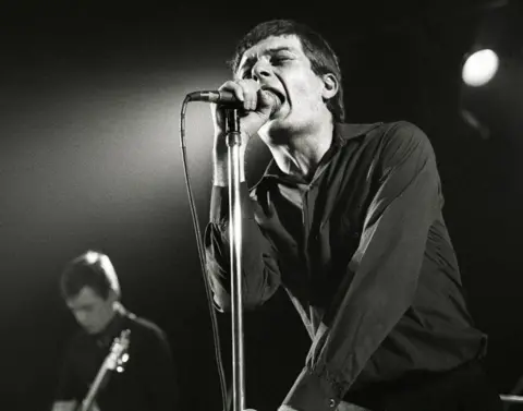 Getty Images Ian Curtis of Joy Division, pictured in 1980, singing into the microphone on stage with his eyes closed.