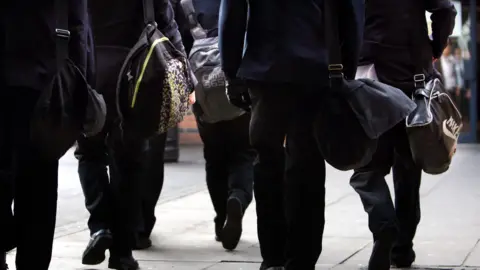 School pupils carrying book bags are walking along a pavement. They were wearing school uniforms.