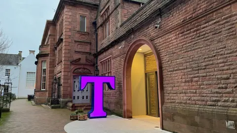 Federica Bedendo/BBC The entrance to Tullie, which has a giant purple illuminate letter T in front of a red-stone building.