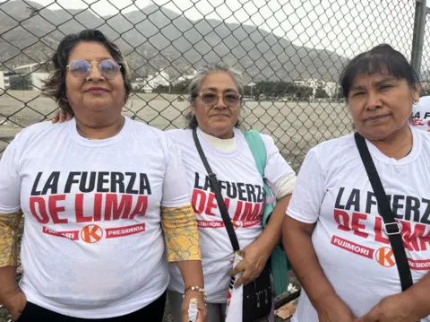 Three women wearing white Fujimori T-shirts, standing in front of a fence with mountains visible behind