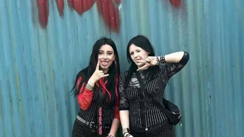 Colby Colby is wearing a red top and a pinstripe waistcoat. She's got her tongue out and is doing a rock and roll hand gesture. She's with her friend who's also wearing pinstripe and doing a peace sign with her tongue out. They're in front of a blue corrugated steel wall.