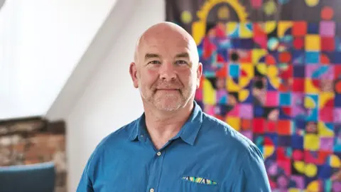 Sue Lambert Trust Clive Evans with very short white hair and beard, wearing a blue shirt and standing in front of a colourful abstract painting or fabric design.