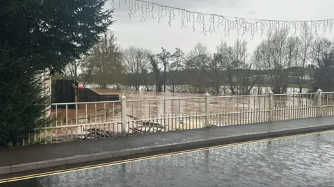 Hereford and Worcester Fire Service A bridge over a river, which is almost as high as the road. There are white iron railings on the bridge's edge, beyond which brown water can be seen, with submerged trees on the river bank. Further flooding can be seen on surrounding fields.