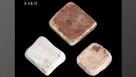 Andrew Williams/Norfolk County Council Three lead ingots against a black background. The top one is roughly square and has a bronzy patina; below on the left is a roughly rectangular ingot and is a whitish shade; below on the right a slightly smaller squarer ingot mottled cream coloured. 
