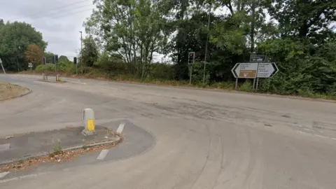 A T-junction on a road with trees in the background and road signs and traffic lights in the distance.