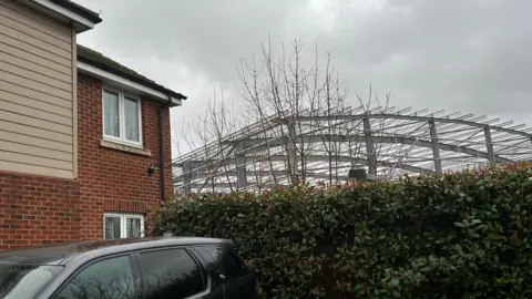 Jo Black/BBC A new two-storey house in a housing estate in the foreground, and the metal frame of a new warehouse building adjacent to the house. There is a hedge separating it from the drive of the house, in which a car is parked.