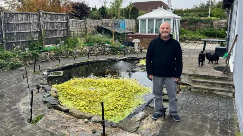 NIGE COOPER Nige Cooper standing beside his pond which has lots of green aquatic plants growing on top of it. The pond has an electric fence surrounding the water and his two chihuahuas are in the back view