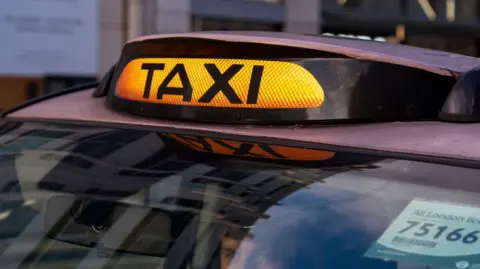 Black taxi with a yellow light on top with the word taxi written on it in black