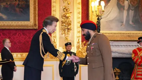Princess Anne shakes hands with Captain Jay Singh-Sohal in a room at St James's Palace. He wears light brown military uniform with a medal on his chest, and a dark blue and red turban. Princess Anne wears a dark skirt suit with gold embellishments. Several men are standing in the background in regal uniforms.