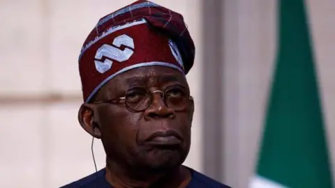 A close-up photo of Nigerian President Tinubu wearing a traditional hat with glasses