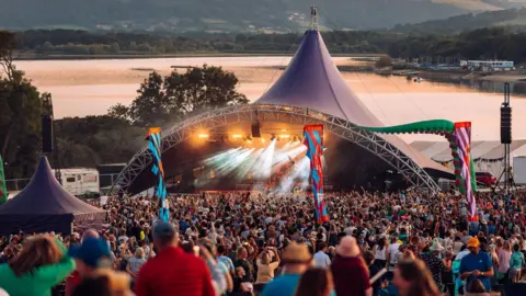 Guilia Spadafora The main tent at Valley Fest, with crowds of people listening to the performers on stage, with banners and the backdrop of Chew Valley Lake.
