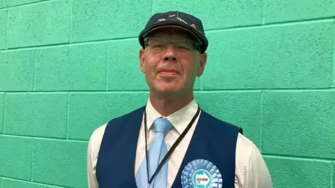 A man in a white shirt with blue waistcoat and blue cap and light blue tie and light blue Reform UK rosette standing against a green wall