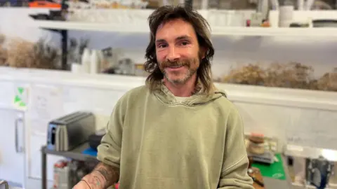 Nathan who has shoulder-length dark hair and a short beard, smiles at the camera behind the counter in his cafe. He is wearing a light green hoodie. 