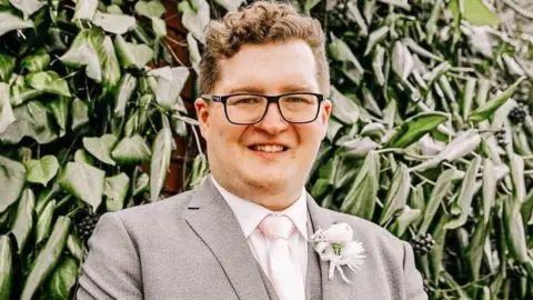 Edward Burr stands smiling at the camera on his wedding day. He wears a light grey suit with a light shirt and tie and a flower in his lapel. He has wavy, brown hair and glasses.