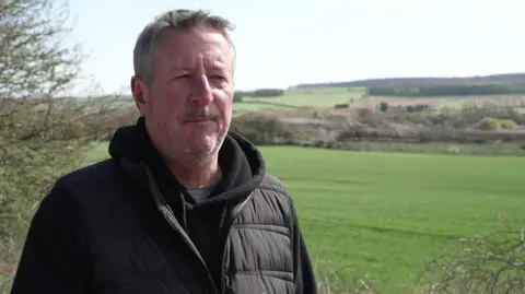A man with short grey hair and a short grey beard stands in front of grassy green field, with a waste management plant in the distance. He wears a black hoodie and a black puffer gilet. 