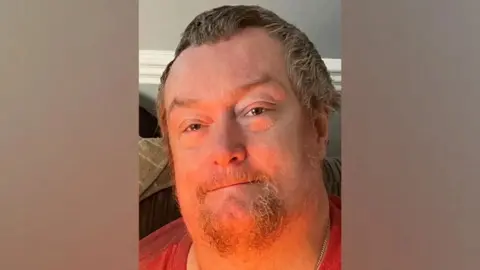 Wayne Ambridge: a man with greying hair and a greying beard wearing a red, round-necked top and smiling 